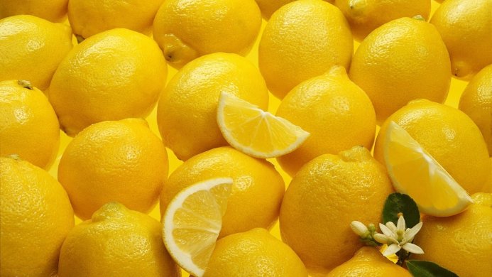 Pile of bright yellow lemons, with lemon slices and a lemon flower on a yellow background