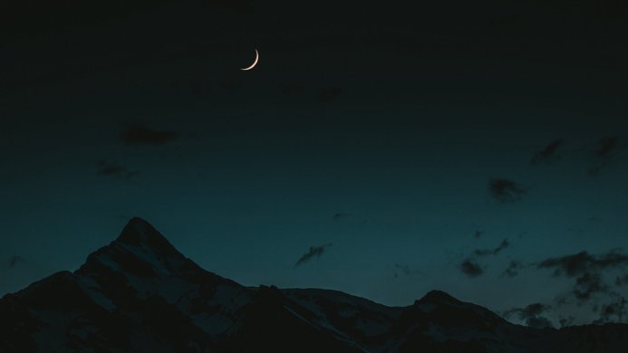 Mountains at night with a crescent moon and sparse clouds in a dark blue sky