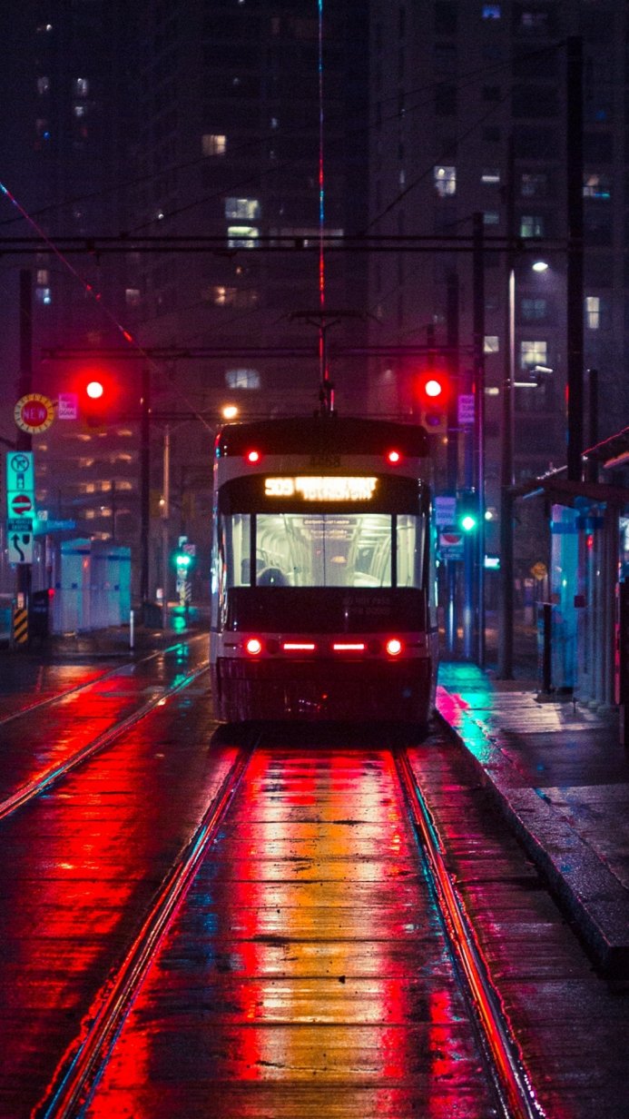 A tram at night on a city street with wet rails reflecting red and blue lights