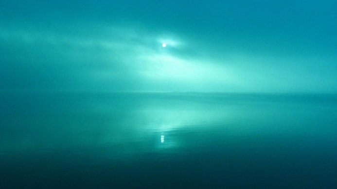 Calm teal sea under a bright sky with the sun shining through the clouds, creating a reflection on the water