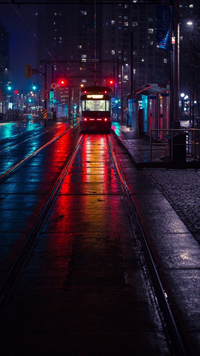A tram driving on a rainy street at night with the reflections of red and blue street lights