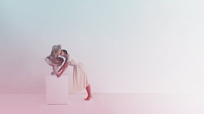 A woman kissing a classical bust sculpture in a pastel-colored room