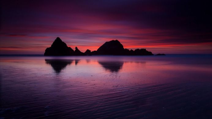 Silhouette of rocks on a beach at sunset with red and purple sky and water reflections
