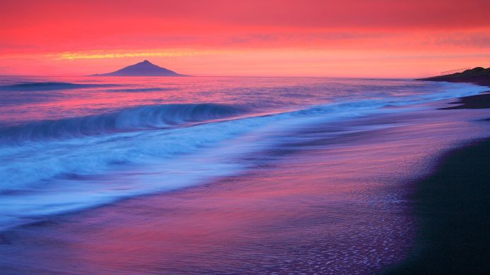 Sunrise on a beach with a mountain in the distance reflecting pink and blue hues