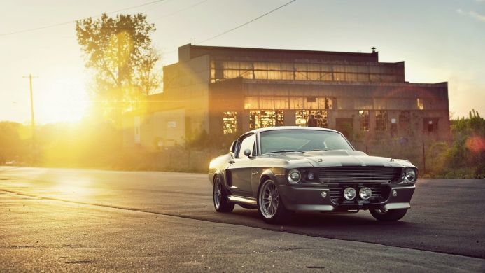 A classic gray Ford Mustang parked in front of an old factory with a golden sunlight