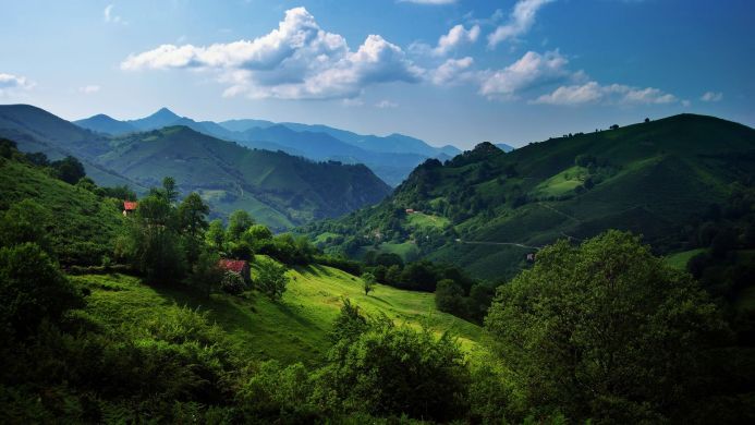 A panoramic view of green mountains with a few small houses and a blue sky with clouds