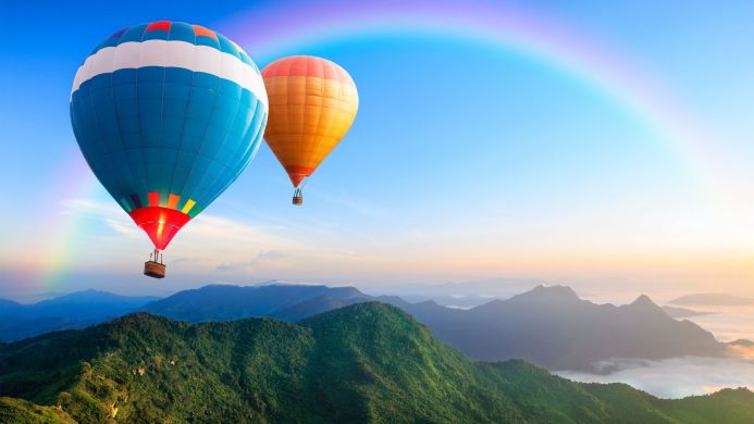 Two hot air balloons over green mountains with a rainbow in the sky