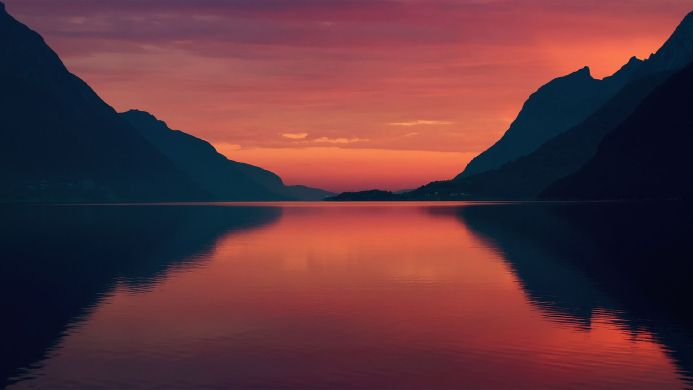 A calm lake reflects the red and orange sky at sunset, surrounded by dark mountains
