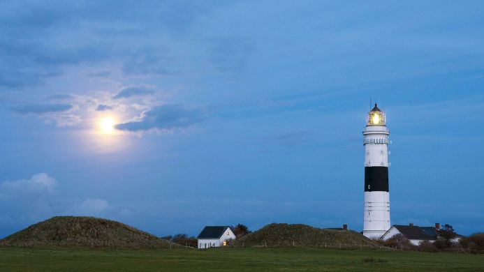 A black and white lighthouse on a coast with a small house under a blue cloudy sky and the moon