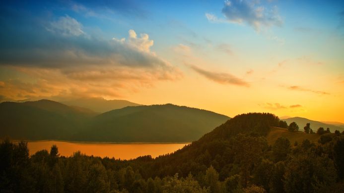 Sunset over mountains, lake, and forest landscape with blue sky and orange sunlight