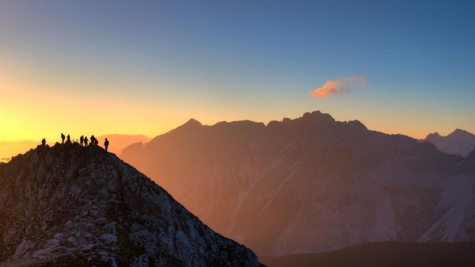 Silhouetted people on a mountain peak at sunset, with mountain range views