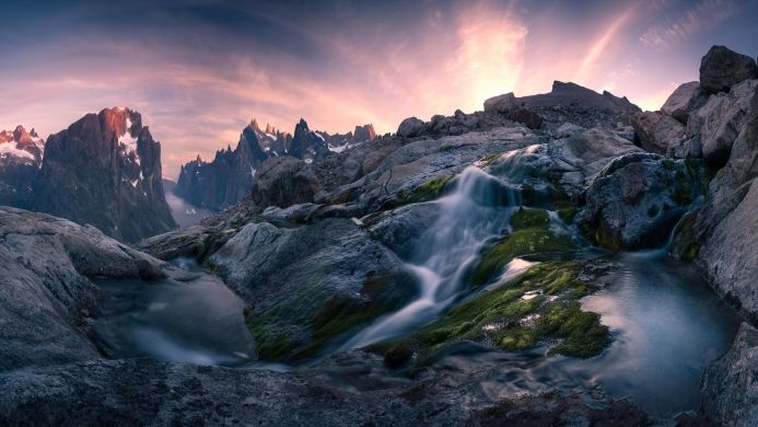 A landscape of a waterfall cascading over mossy rocks with mountains in the background under a sunset sky