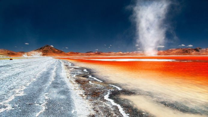 A red lake with a dust devil in a mountainous arid landscape under a dark blue sky