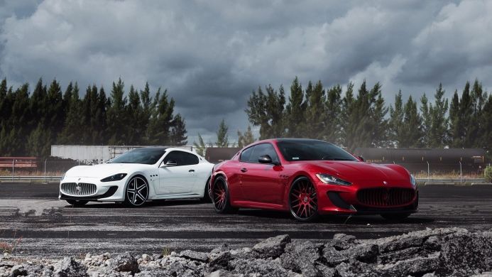 Two Maserati GranTurismo cars, one red and one white, on a gray asphalt with an overcast sky