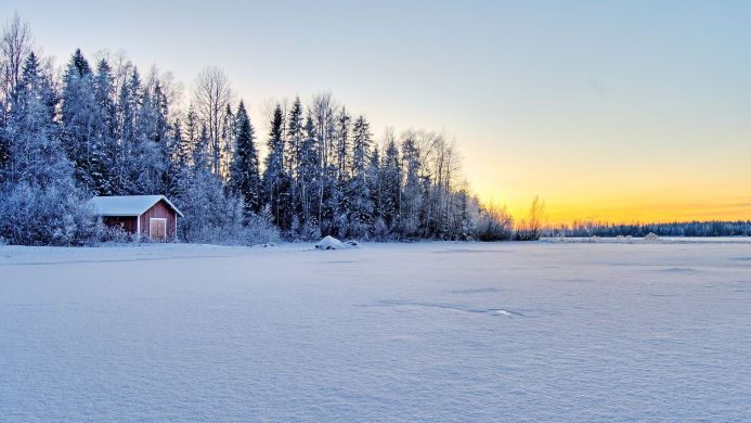 Snow covered cabin next to a frozen lake surrounded by trees in winter at sunset