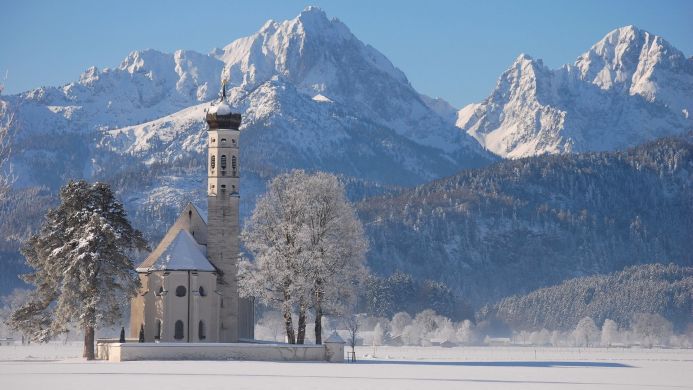 A white church stands in a snow-covered field with mountains in the background