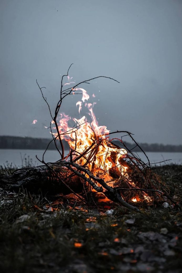 A crackling bonfire burns brightly against a gray sky near a body of water