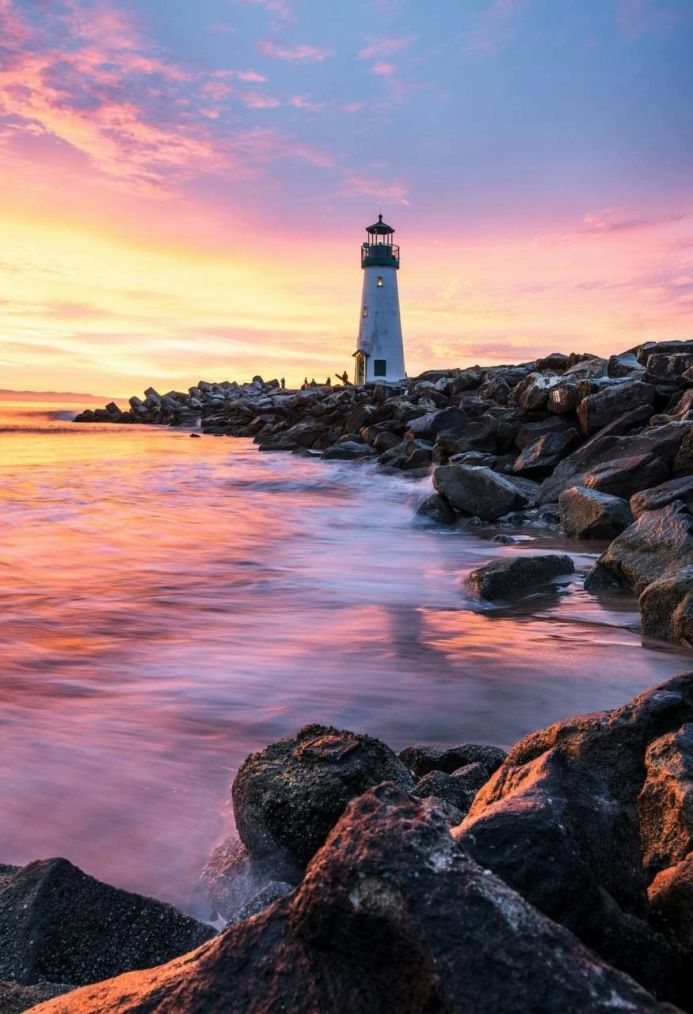 A lighthouse stands on a rocky shore during a colorful sunrise or sunset
