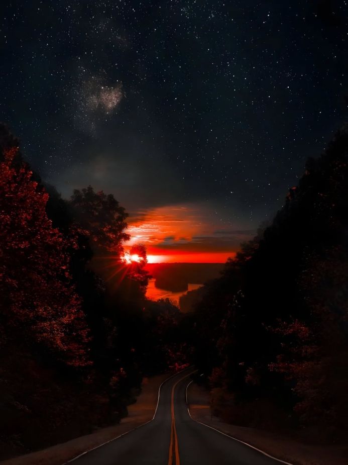 Road leading to a fiery sunset under a starry sky, framed by trees