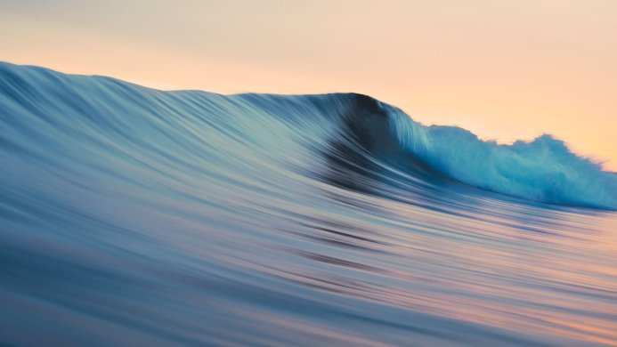 A large blue wave cresting in the ocean with a peach-colored sky