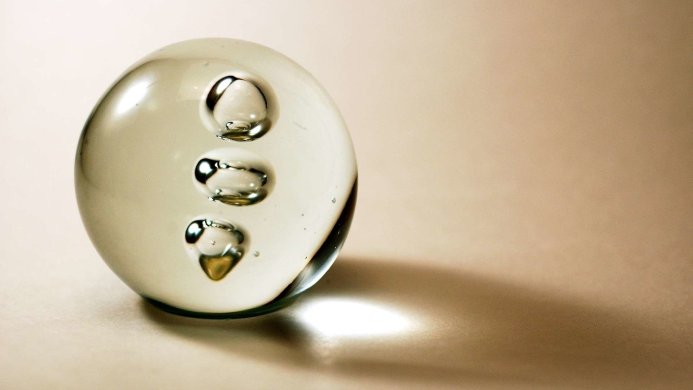 A clear glass sphere with bubbles inside on a beige surface, casting a soft shadow