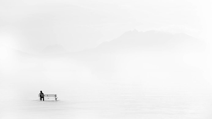 A person sits on a bench, looking out at a misty mountain landscape in monochrome