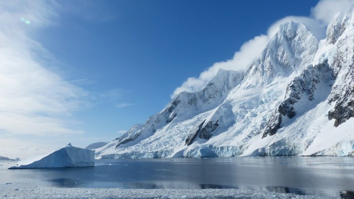 An iceberg floats in the water near snow-covered mountains under a blue sky in Antarctica