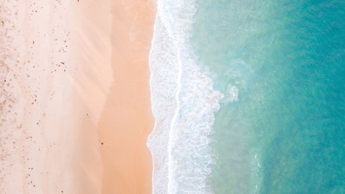 Aerial view of a beach with turquoise water and beige sand with white waves breaking on the shore