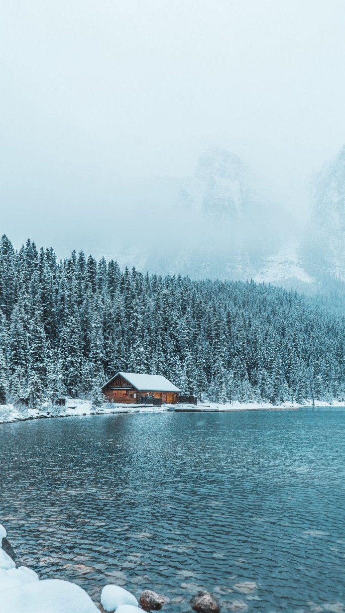 A cabin by a lake surrounded by snow-covered trees and mountains in winter