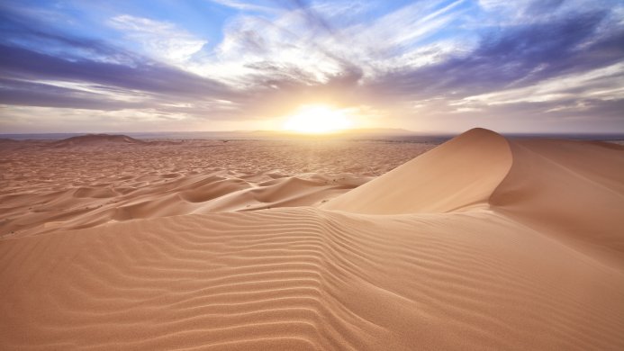A large sand dune in a desert landscape with a bright sun rising over the horizon