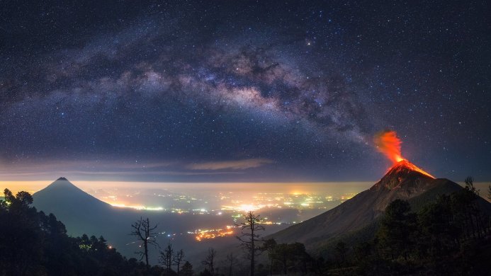 Two volcanoes, one erupting, under the Milky Way with a city glowing in the valley below
