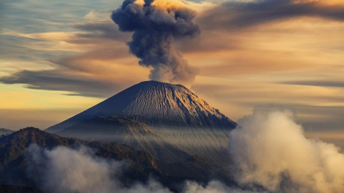 A volcano erupts, sending smoke and ash into the sky during a sunrise or sunset, with clouds in the foreground