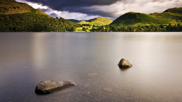 A peaceful lake surrounded by mountains and green trees with two stones in the water