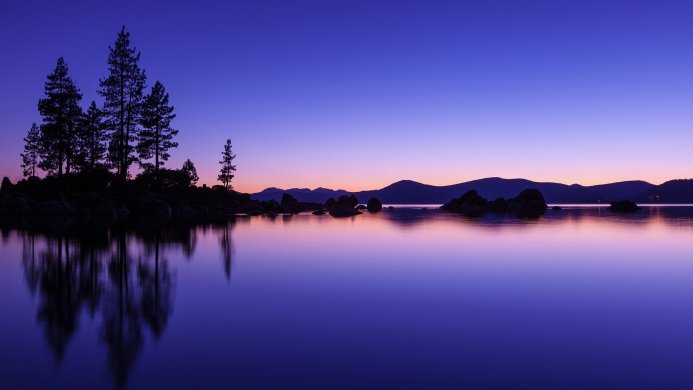 Lake Tahoe at sunset with trees, rocks and mountains reflecting in the still water