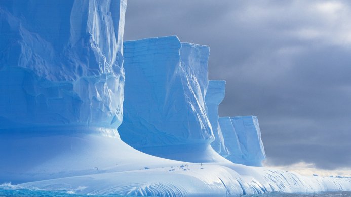 Large iceberg with textured blue ice and a few penguins on top against a cloudy sky