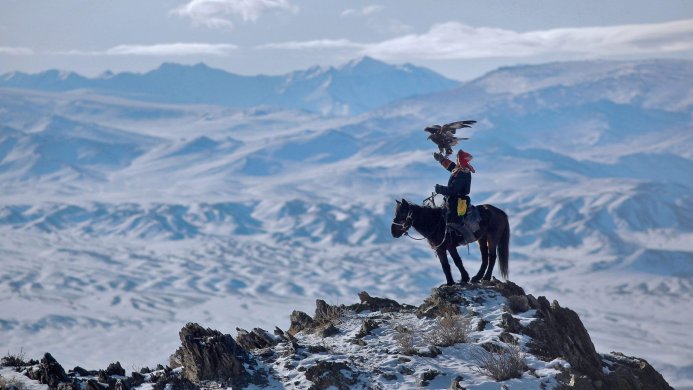 Mongolian eagle hunter on horseback with his golden eagle in a snow-covered mountain landscape