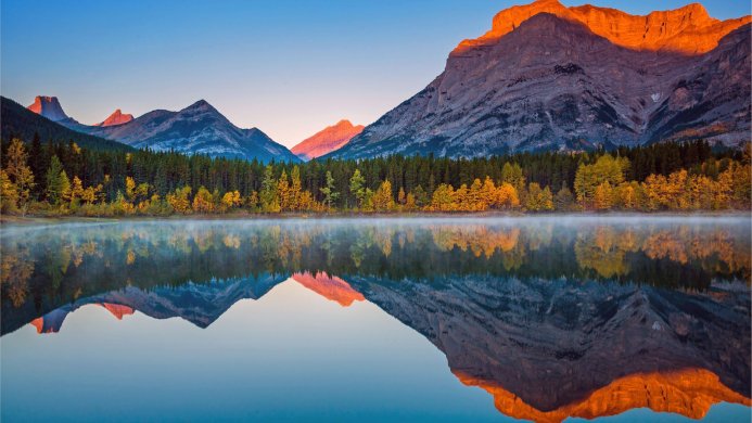 Mountain range reflected in a calm lake with trees, orange light on peaks