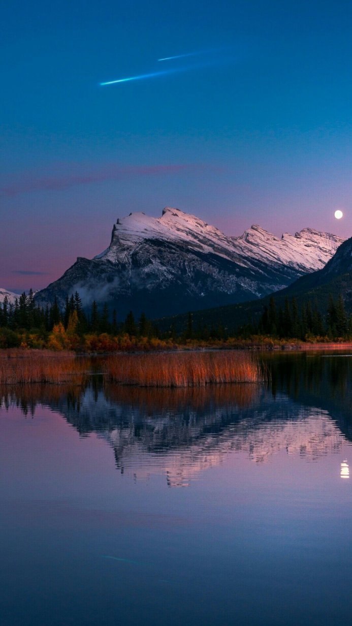 Snowy mountain reflecting in a lake under a twilight sky with a shooting star and the moon