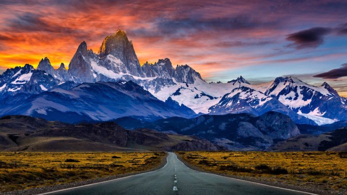A road leading to snow-capped mountains under an orange and blue sky