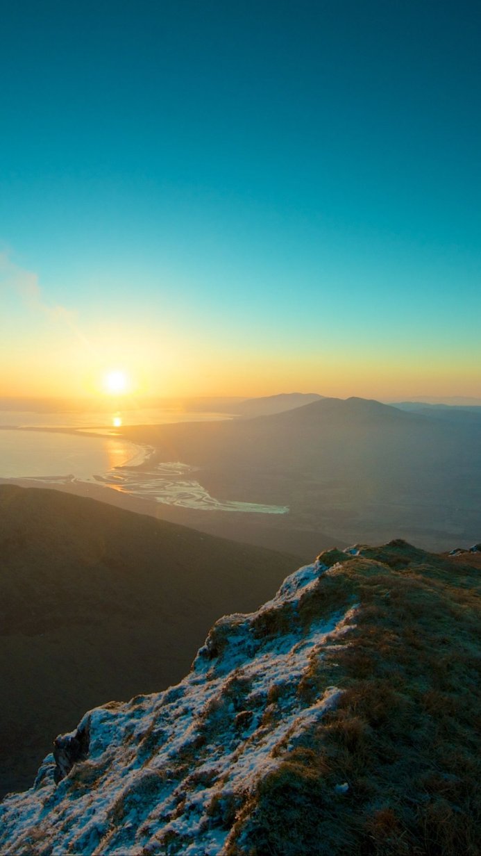 Sunrise over snow-capped mountains near a coastline, with a gradient of blue and gold in the sky