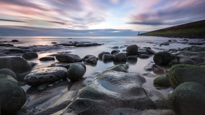 Rocky beach at dusk with a lighthouse in the distance and smooth, long-exposure water