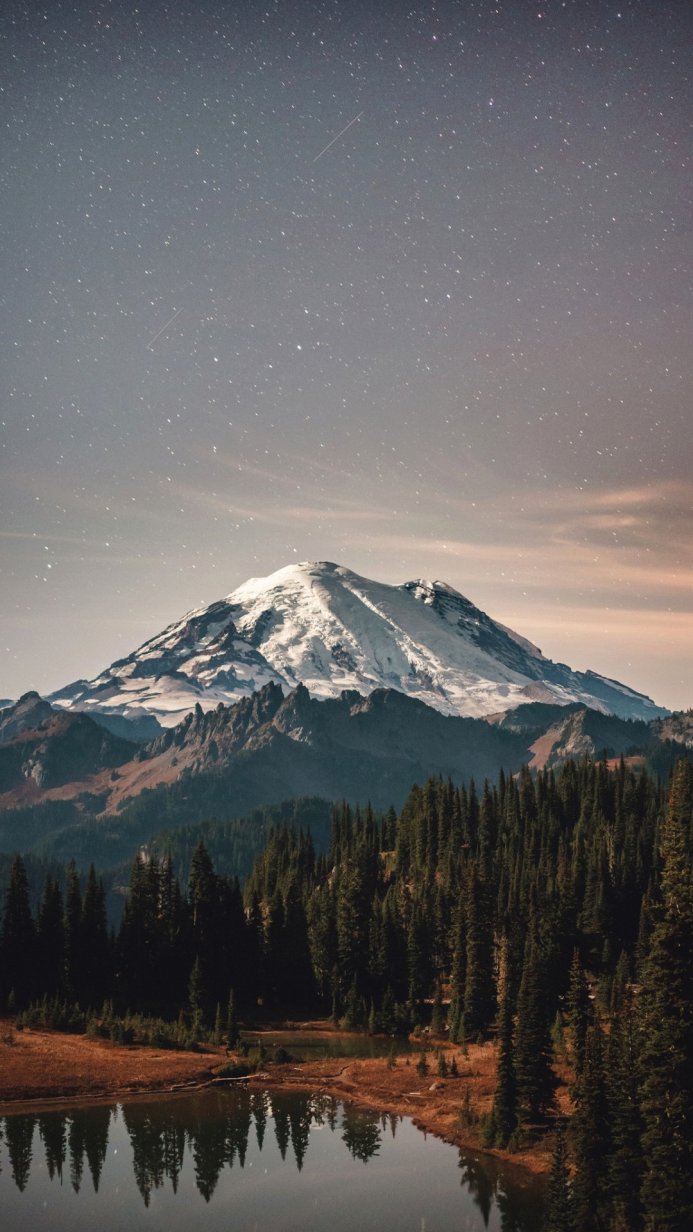 Snowy mountain peak under a starry night sky reflected in a lake