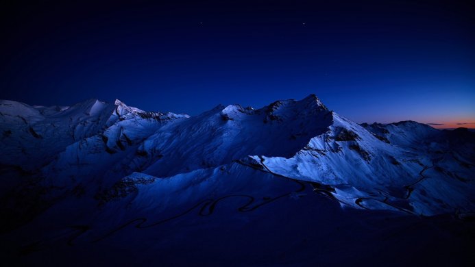 Snowy mountain range at night under a dark blue sky with visible stars and a winding road