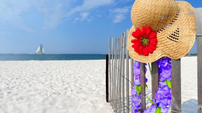 Straw hat and flowers on a fence with a beach and sailboat in the background