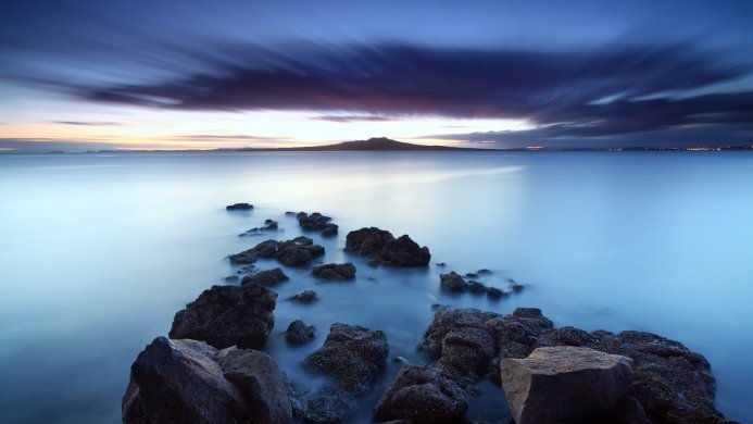 A tranquil seascape with rocks in the foreground and a distant mountain under a colorful twilight sky