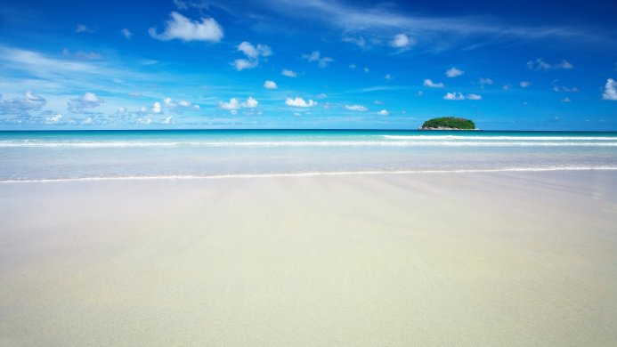 A tropical island sits off a white sand beach under a blue sky with fluffy white clouds