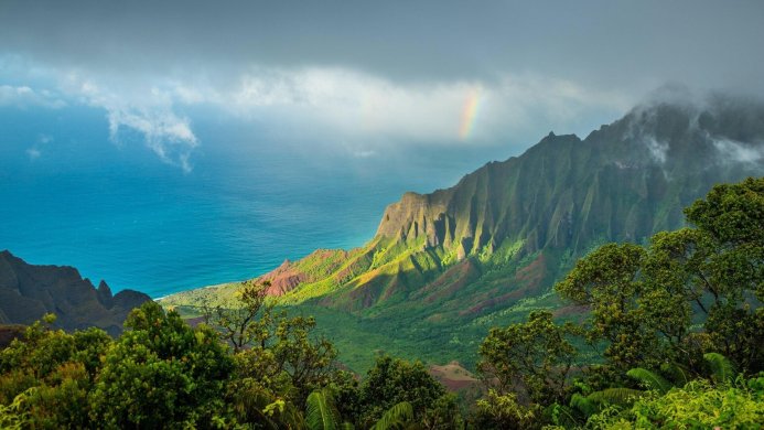 A scenic view of green mountains meeting the blue ocean, with trees in the foreground and a rainbow in the background