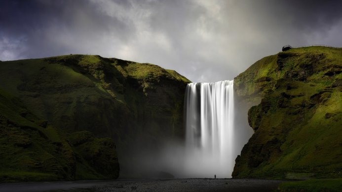 A long exposure shot of a powerful waterfall flowing between green hills under a cloudy sky