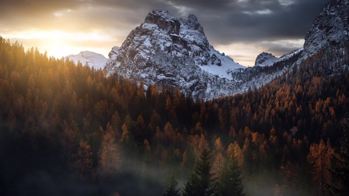Snow-covered mountain peak surrounded by an orange forest, with a cloudy sky