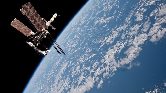 The International Space Station orbiting Earth with a blue sky and white clouds in the background